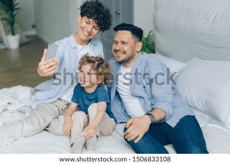 Perfect young family girl guy and kid taking selfie at home in bed using smartphone camera posing smiling. Photograph, happiness and technology concept.