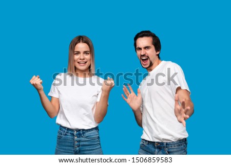 Excited couple expressing emotion of success. Young blond woman and bearded man with mustaches in white tees and blue jeans are winner. Models standing isolated over blue background.