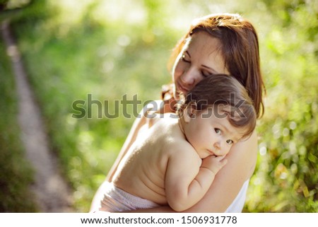 A child in her mother’s arms. She pressed herself against her shoulder. She laid her head on her mother, calmly looking at the camera. Tender photo. Natural light. The joy and tenderness of motherhood
