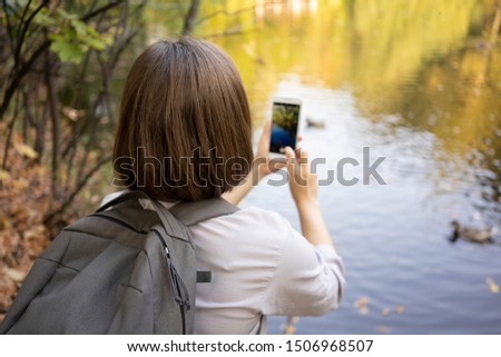 Woman taking a picture of ducks in lake while walking in autumn park with a smart phone