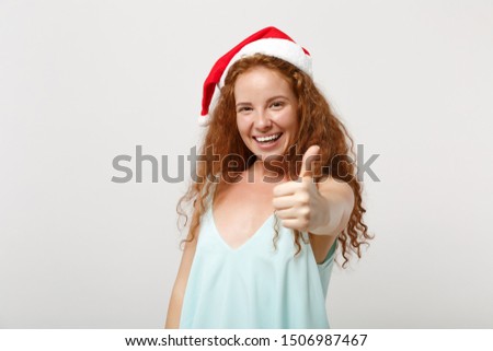Joyful young redhead Santa girl in light clothes, Christmas hat isolated on white background, studio portrait. Happy New Year 2020 celebration holiday concept. Mock up copy space. Showing thumb up