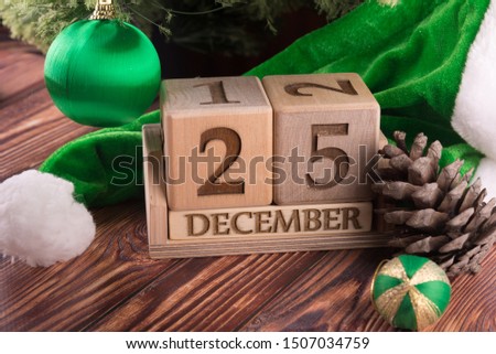 New year, Christmas composition. Wooden cube calendar with 25 december date staying on table near green Santa hat, pine tree cone and decorative balls on brown table