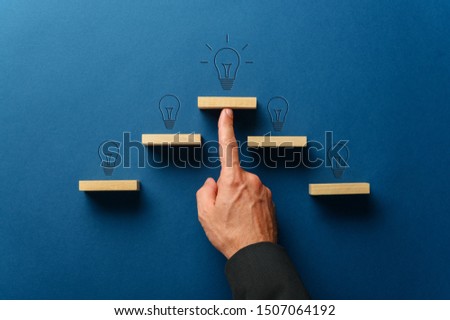 Male finger supporting the top wooden peg in a pyramid shaped stairway  with a light bulb on top of every step . Over navy blue background.