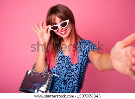 Pleasant girl with pink hair making selfie in studio and laughing. Good-looking young woman in blue shirt and bag taking picture of herself on bright pink background.