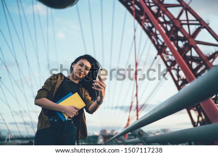 Attractive female influencer in optical eyewear photographing herself via front cellular camera posing at urban setting, beautiful hipster girl with smartphone device making decent media content
