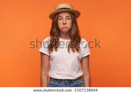 Studio photo of cute red haired young woman posing over orange background with upset face, looking to camera and grimacing, posing over orange background