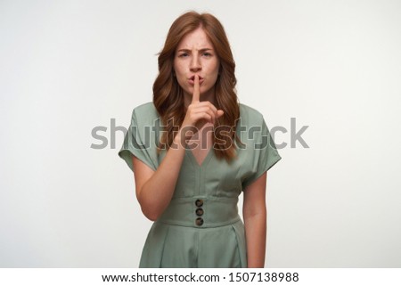 Serious red haired young woman in romantic dress looking to camera with raised forefinger to her mouth, making hush gesture over white background