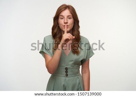 Portrait of beautiful young redhead female in vintage pastel dress posing over white background, raising index finger to lips, asking to keep secret