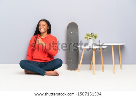African American skater teenager girl with braided hair sitting on the floor giving a thumbs up gesture