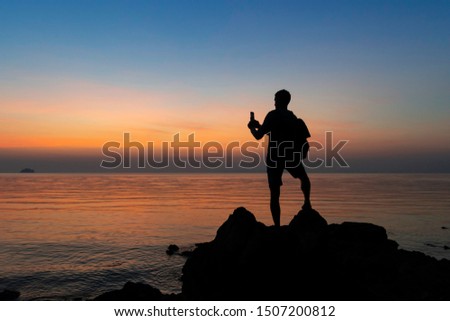 Silhouette of a man with a backpack standing on the rock by the sea at sunset and taking pictures on a smartphone 