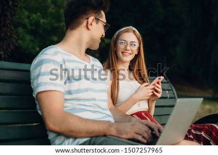 Young red haired girl with freckles using a smartphone while looking at her boyfriend. Two young student sitting on a bench.