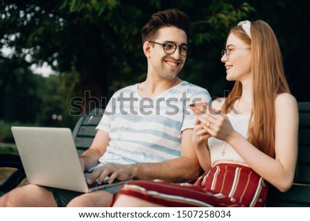 Happy young couple working while sitting on a bench in the park. Young programmer typing data code while sitting with his girlfriend.