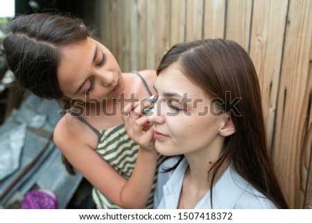 Little girl learn to do a makeup on her older sister, applying liquid tonal foundation on the face. Outdoor photo on wood wall background. Summer holidays