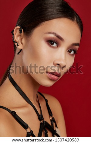 Cropped side view shot of woman with straight dark hair with side part and ponytail. The girl with carmine colour lips in black bra-harness is wearing black cuff earring in shape of a snake. 