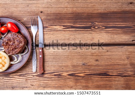 Fresh grilled beefsteaks with vegetables - onions and tomatoes - on brown vintage style plate on wooden background with fork and knife. Grilled food. Copy space.