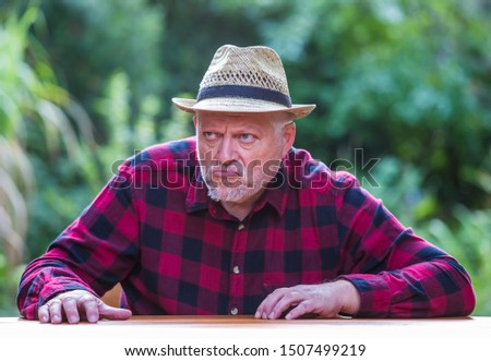 An elderly man in a hat is sitting with bad mood at a table in the garden. Concept: evil and unsympathetic people Royalty-Free Stock Photo #1507499219