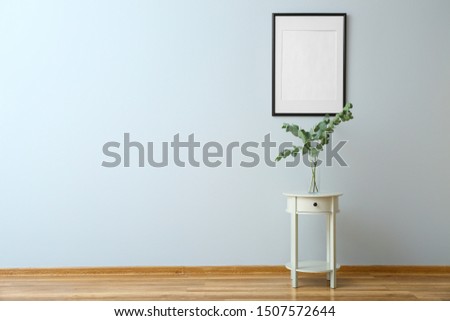 Table with eucalyptus branches in vase and frame hanging on light wall