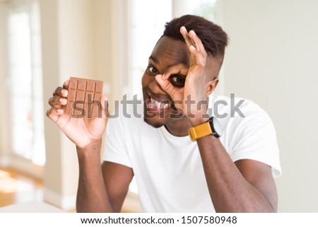 Young african american man eating chocolate bar with happy face smiling doing ok sign with hand on eye looking through fingers