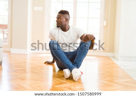 Handsome african american man sitting on the floor at home looking to side, relax profile pose with natural face with confident smile.