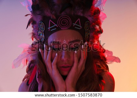 Portrait of thinking nice woman in headdress of feathers propping her head and looking downward isolated over violet neon background