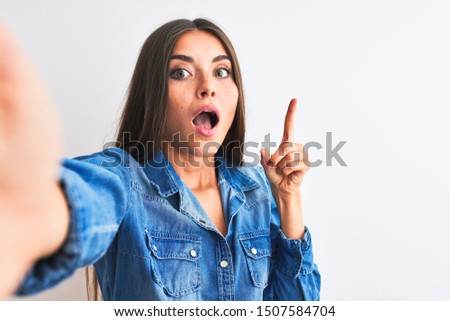 Beautiful woman wearing denim shirt make selfie by camera over isolated white background pointing finger up with successful idea. Exited and happy. Number one.