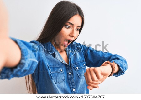 Beautiful woman wearing denim shirt make selfie by camera over isolated white background Looking at the watch time worried, afraid of getting late