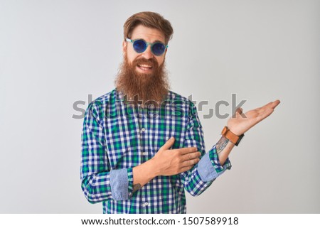 Young redhead irish man wearing casual shirt and sunglasses over isolated white background smiling cheerful presenting and pointing with palm of hand looking at the camera.