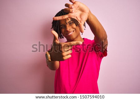 Afro american man with dreadlocks wearing t-shirt standing over isolated pink background smiling making frame with hands and fingers with happy face. Creativity and photography concept.