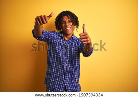 Afro man with dreadlocks wearing casual shirt standing over isolated yellow background approving doing positive gesture with hand, thumbs up smiling and happy for success. Winner gesture.