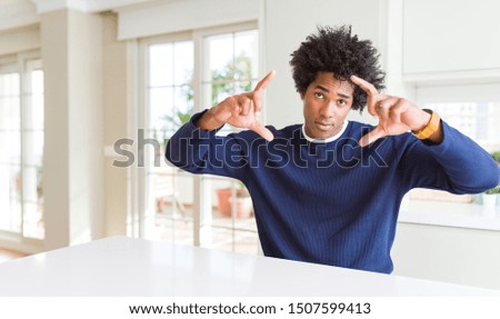 Young african american man wearing casual sweater sitting at home doing frame using hands palms and fingers, camera perspective