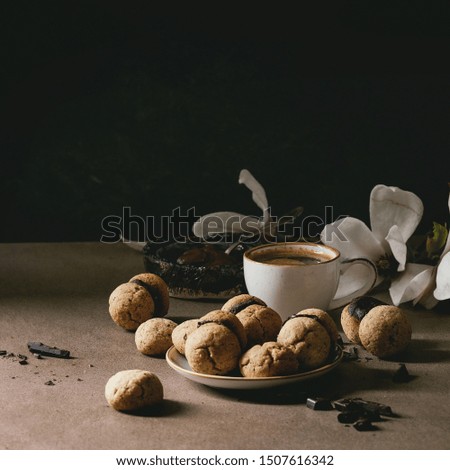 Baci di dama homemade italian hazelnut biscuits cookies with chocolate cream served in ceramic plate with cup of espresso coffee and magnolia flowers on brown table. Dark still life.