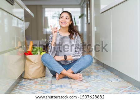 Young woman sitting on the kitchen floor with a paper bag full of fresh groceries with a big smile on face, pointing with hand and finger to the side looking at the camera.