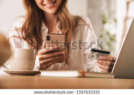 Cropped photo of young cheerful ginger business woman sit indoors in office using laptop computer and mobile phone holding credit card.