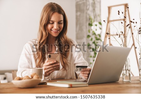 Photo of young happy redhead business woman sit indoors in office using laptop computer and mobile phone holding credit card.