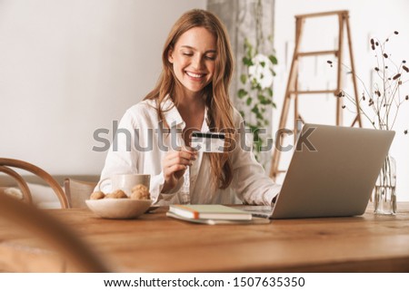 Image of young cheerful ginger business woman sit indoors in office using laptop computer holding credit card.