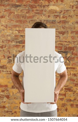 Front view close up of a young Caucasian man holding a blank white canvas in his hands which covers his face, standing in front of a brick wall