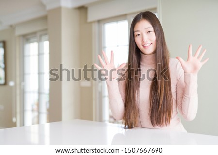 Beautiful Asian woman wearing casual sweater on white table showing and pointing up with fingers number ten while smiling confident and happy.