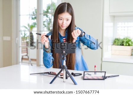 Beautiful Asian influencer woman recording make up tutorial using smartphone camera with angry face, negative sign showing dislike with thumbs down, rejection concept