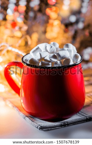 Red mug with hot chocolate and a lot of little marshmallows. Christmas lights in the background.