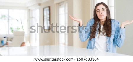 Wide angle picture of beautiful young woman sitting on white table at home clueless and confused expression with arms and hands raised. Doubt concept.