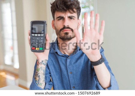 Young man holding dataphone point of sale as payment with open hand doing stop sign with serious and confident expression, defense gesture