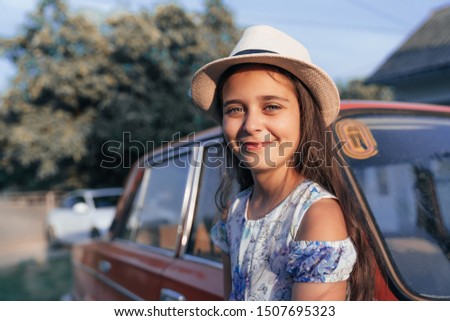 Clsose up portrait, beautiful young brunette little girl in dress and hat leaned on the red old antique car and looking at the caamera, summer time