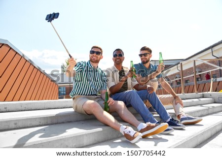 leisure, technology and people concept - happy male friends taking picture by smartphone on selfie stick and drinking beer on street in summer