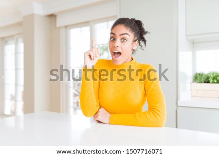 Beautiful african american woman with afro hair wearing a casual yellow sweater pointing finger up with successful idea. Exited and happy. Number one.