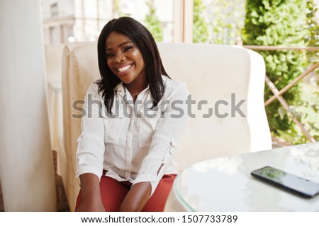 Formally dressed african american business woman in white blouse and red trousers sitting on outdoor terrace. Successful dark skinned businesswoman.
