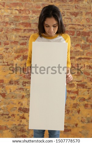 Front view close up of a young Caucasian woman holding a blank white canvas in her hands and flooking down at it, standing in front of a brick wall
