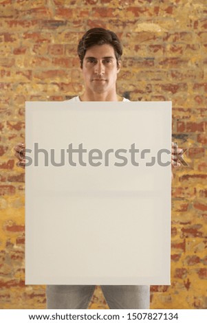 Front view close up of a young Caucasian man holding a blank white canvas in his hands and facing camera, standing in front of a brick wall