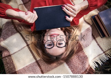 Young blond woman, wearing eyeglasses and red shirt, lying on brown blanket, holding black old book in her hands. Close-up picture of pretty student, relaxing after studies, Creative flat-lay portrait
