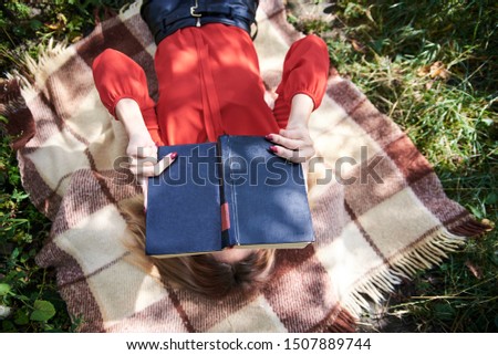 Young blond woman, wearing eyeglasses and red shirt, lying on brown blanket, covering her face with black old book in her hands. Close-up picture of pretty student, relaxing after studies. College fun
