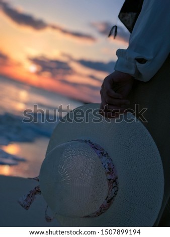 A person is holding a hat while enjoying the sunrise at the beach.